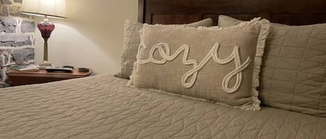 A bed with a quilted bedspread and two pillows, one with a 'lozy' embroidered design. A lamp with a red shade and a book are on a wooden nightstand beside the bed.