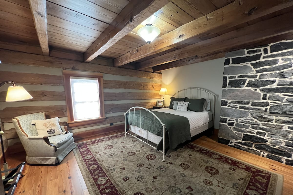 A cozy bedroom with a stone wall, wooden ceiling beams, a metal bed frame, a chair with a cushion, a bedside table with a lamp, and a patterned rug.
