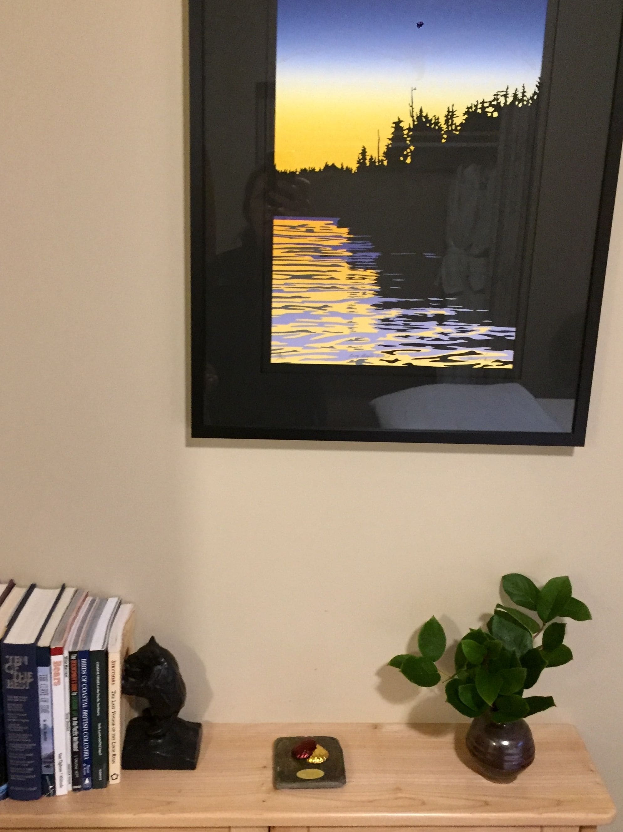 A framed picture of a sunset over water, a bookshelf with books, a small plant, and a decorative item on a wooden shelf.