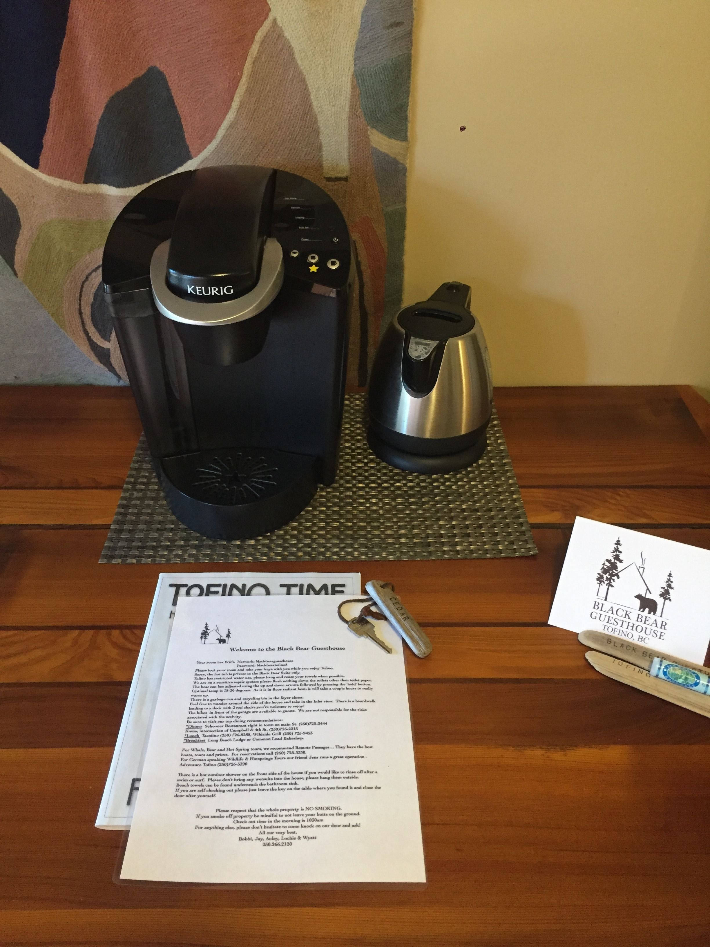 A Keurig coffee maker, a kettle, a welcome sign, and a set of keys on a wooden table.