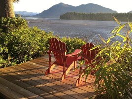 Una terraza de madera con dos sillas Adirondack rojas que dan a un cuerpo de agua y montañas al fondo.