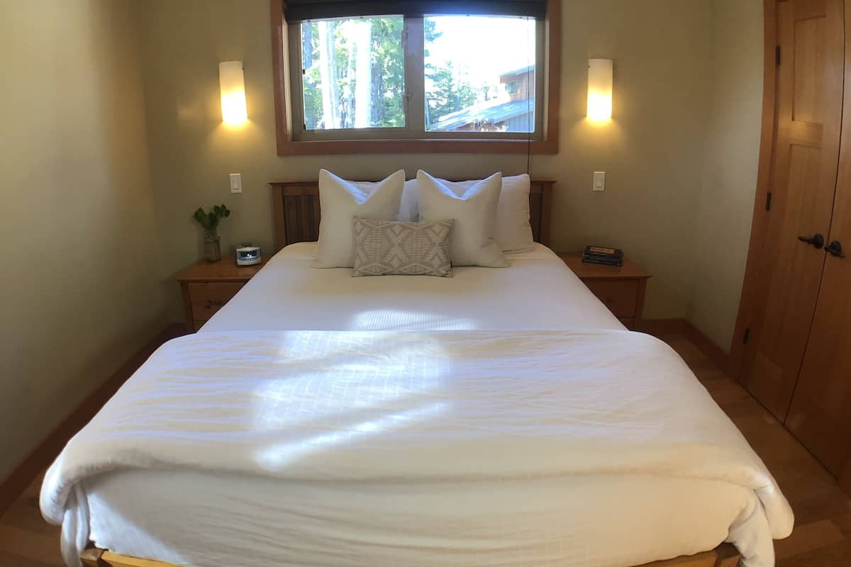 Een groot bed met houten frame, twee nachtkastjes met lampen, een raam met uitzicht op bomen en een houten deur.