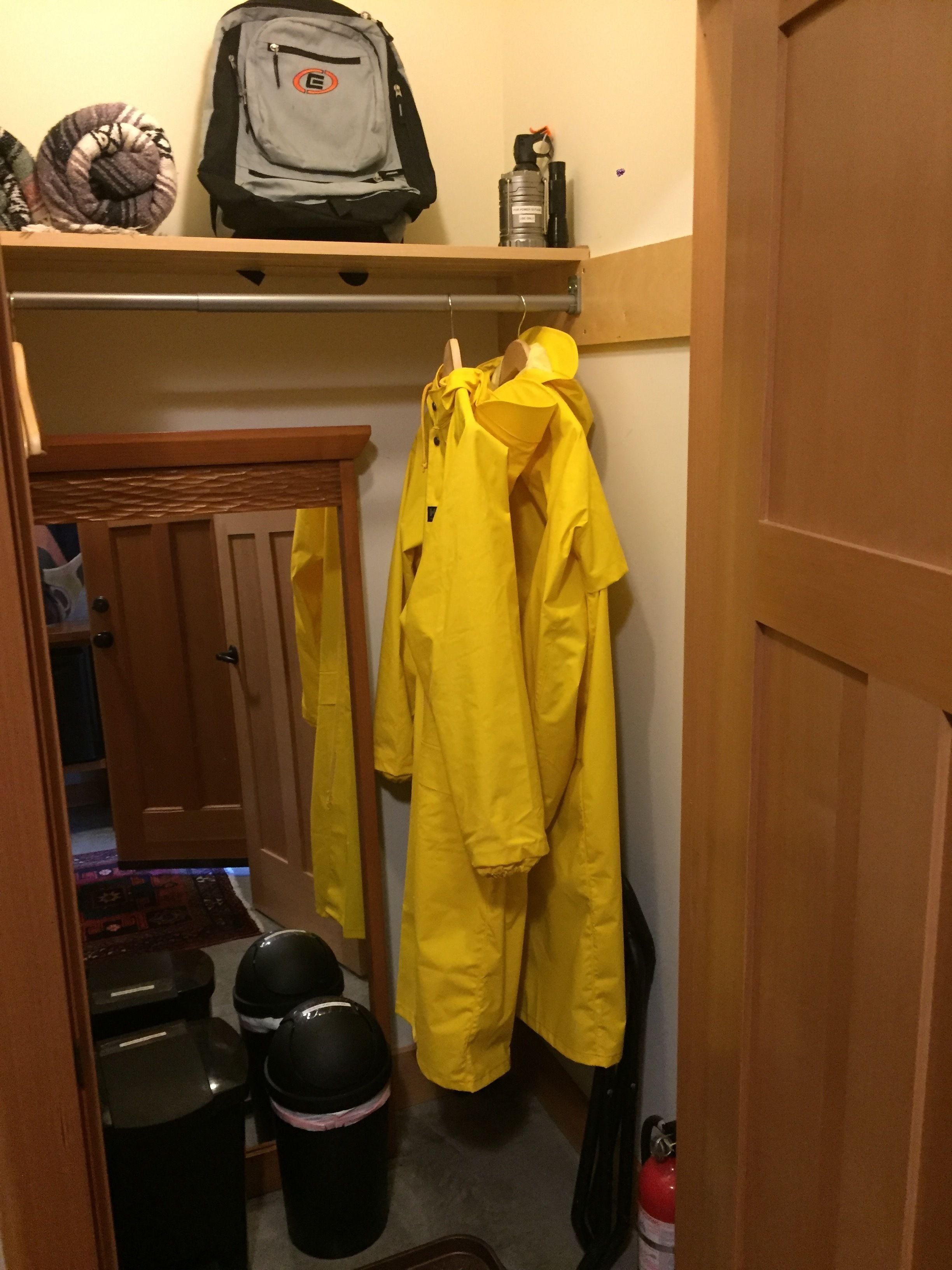Un petit placard avec une porte en bois, une étagère sur laquelle se trouvent un sac à dos et une bouteille d’eau, et une paire de vestes de pluie jaunes accrochées à la porte.