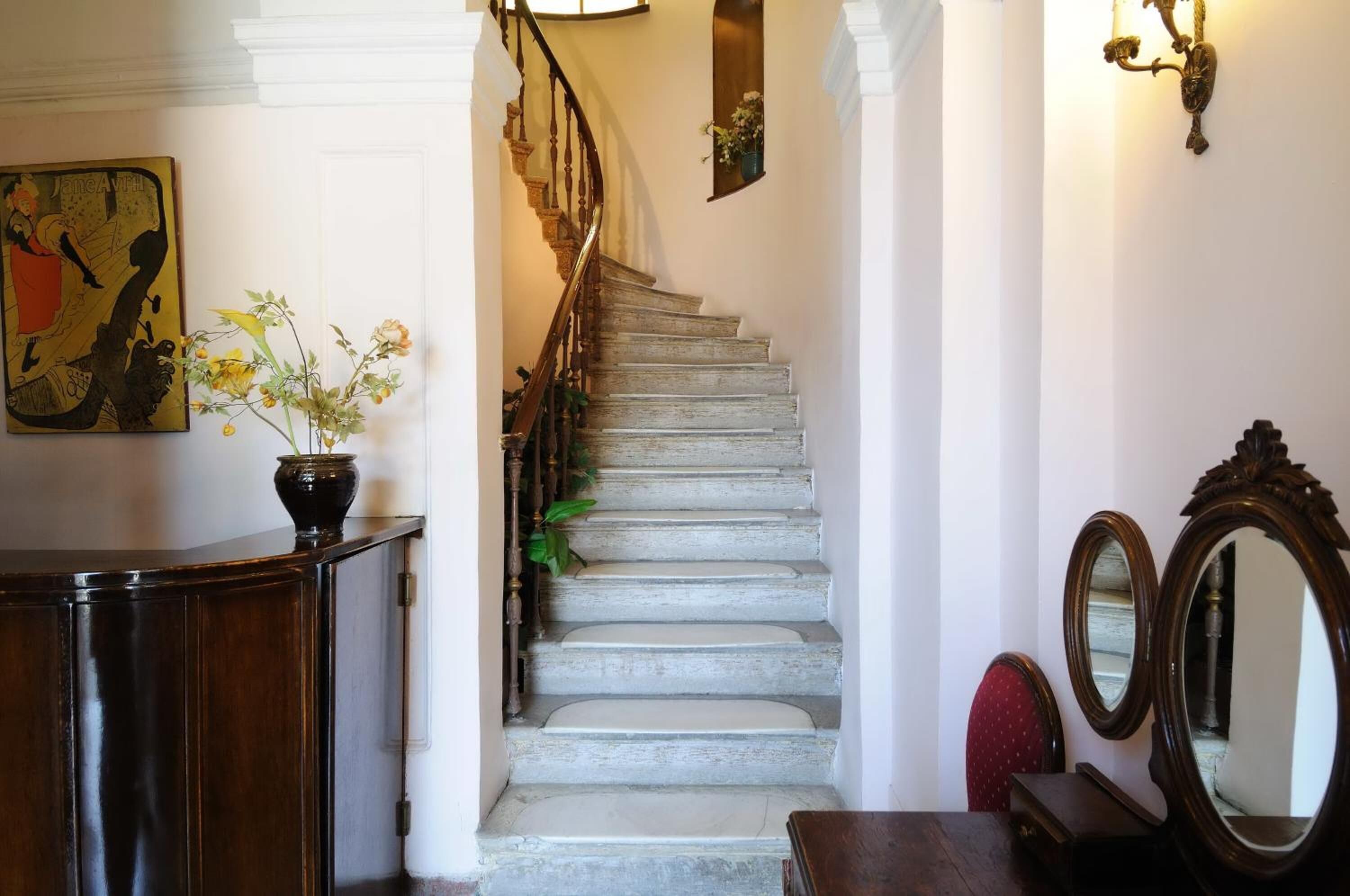 Un escalier avec une rampe en bois et un mur blanc. Un vase rempli de fleurs se trouve à gauche et un miroir à droite.