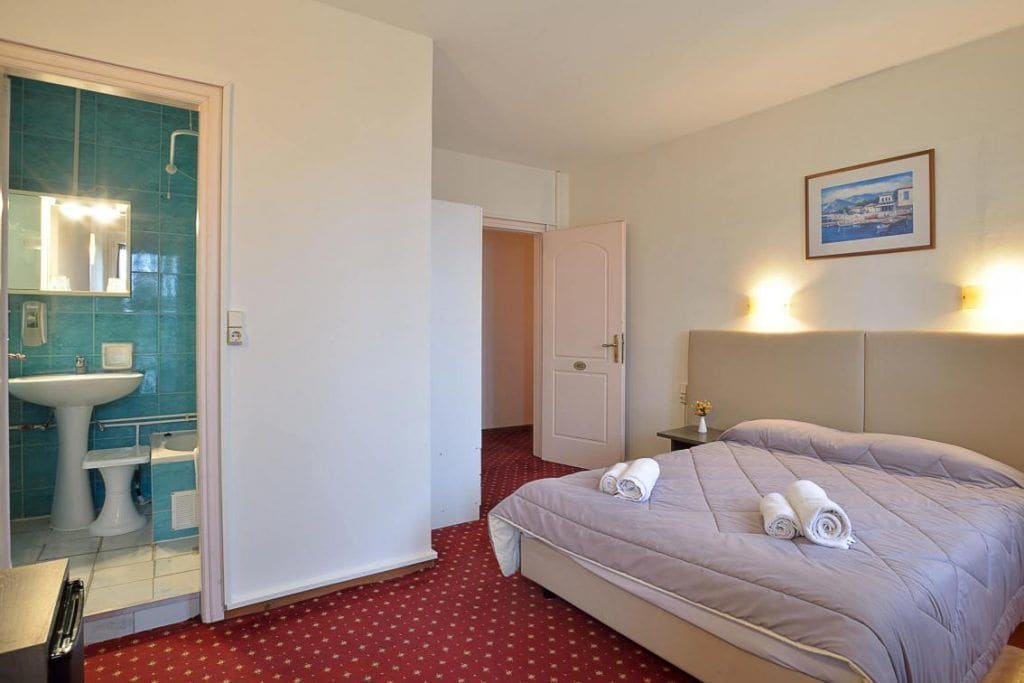 Une chambre d’hôtel comprenant un lit, une salle de bain avec un lavabo et un miroir, et une porte donnant sur une autre pièce.