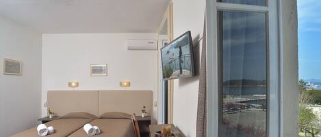 Habitación de hotel con dos camas, un televisor, un ventanal con vista al puerto y una pequeña mesa con un florero.