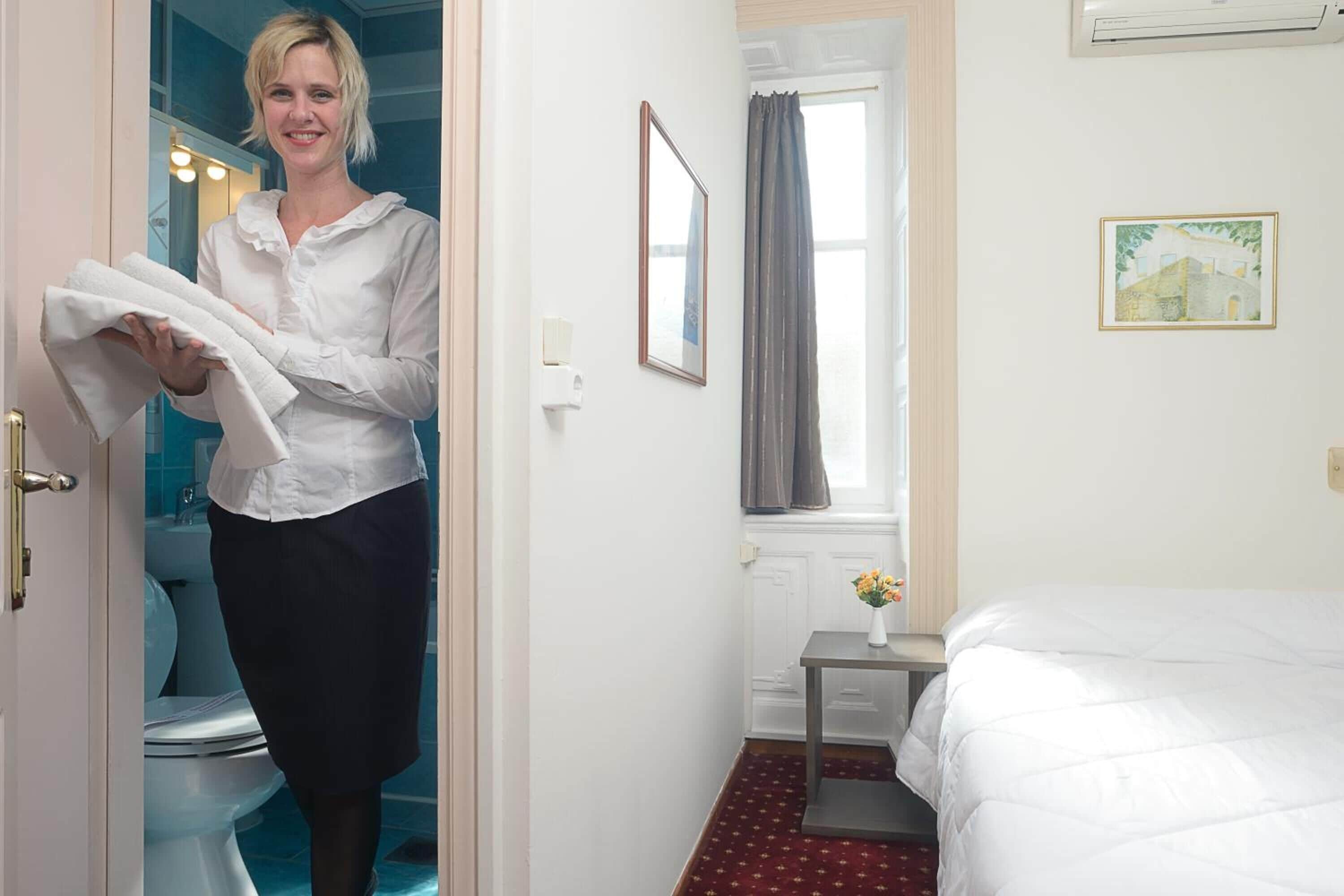 Ein Hotelzimmer mit Bett, einem kleinen Tisch mit Blumenvase, einem Fenster mit Vorhängen und einem gerahmten Bild an der Wand.