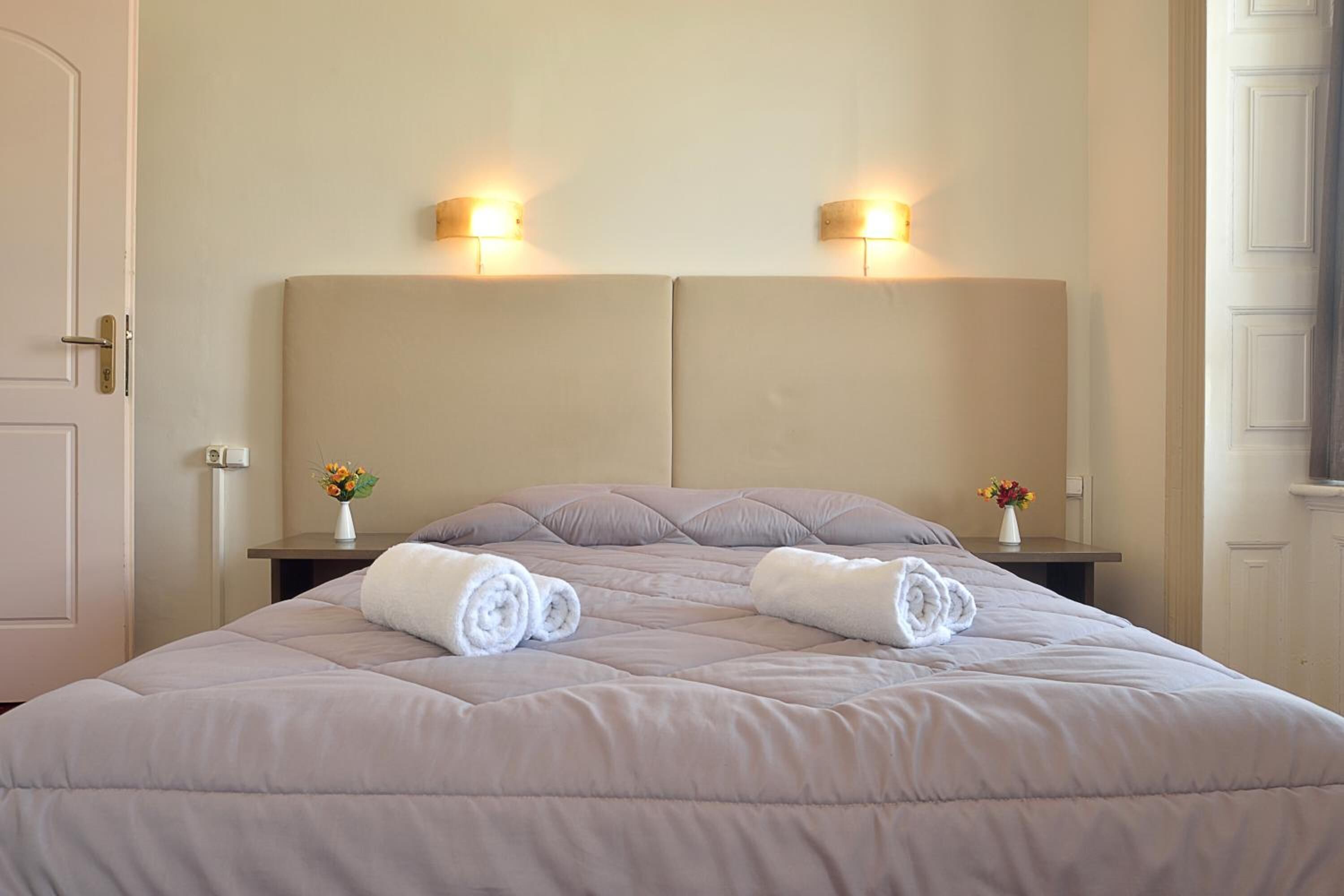 Een netjes opgemaakt bed met een gewatteerd hoofdbord, twee nachtkastjes met lampen, en een vaas met bloemen aan beide zijden.