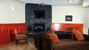 A living room with a stone fireplace, a flat-screen TV mounted above it, a leather sofa with orange cushions, a patterned armchair, and a framed photo on the wall. - Trend Mountain Hotel & Conference Centre (Tumbler Ridge)
