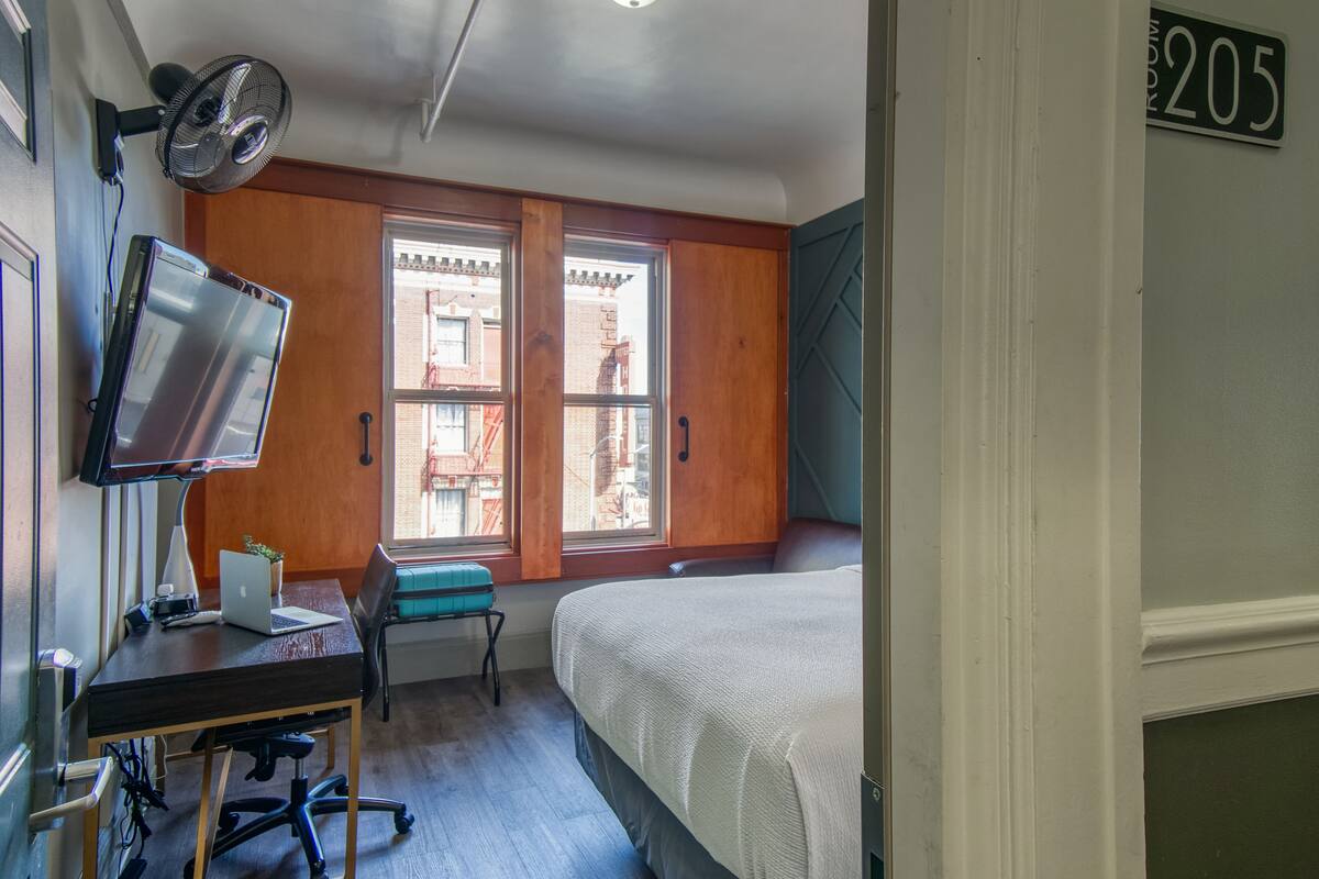 A room with a bed, a desk with a laptop, a chair, a standing fan, and a window with a view of buildings.