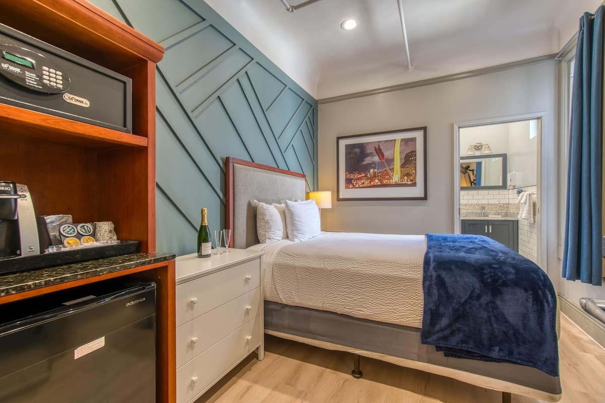 A hotel room with a bed, a nightstand, a minibar, a wall-mounted TV, a framed picture, and a bathroom visible through an open door.