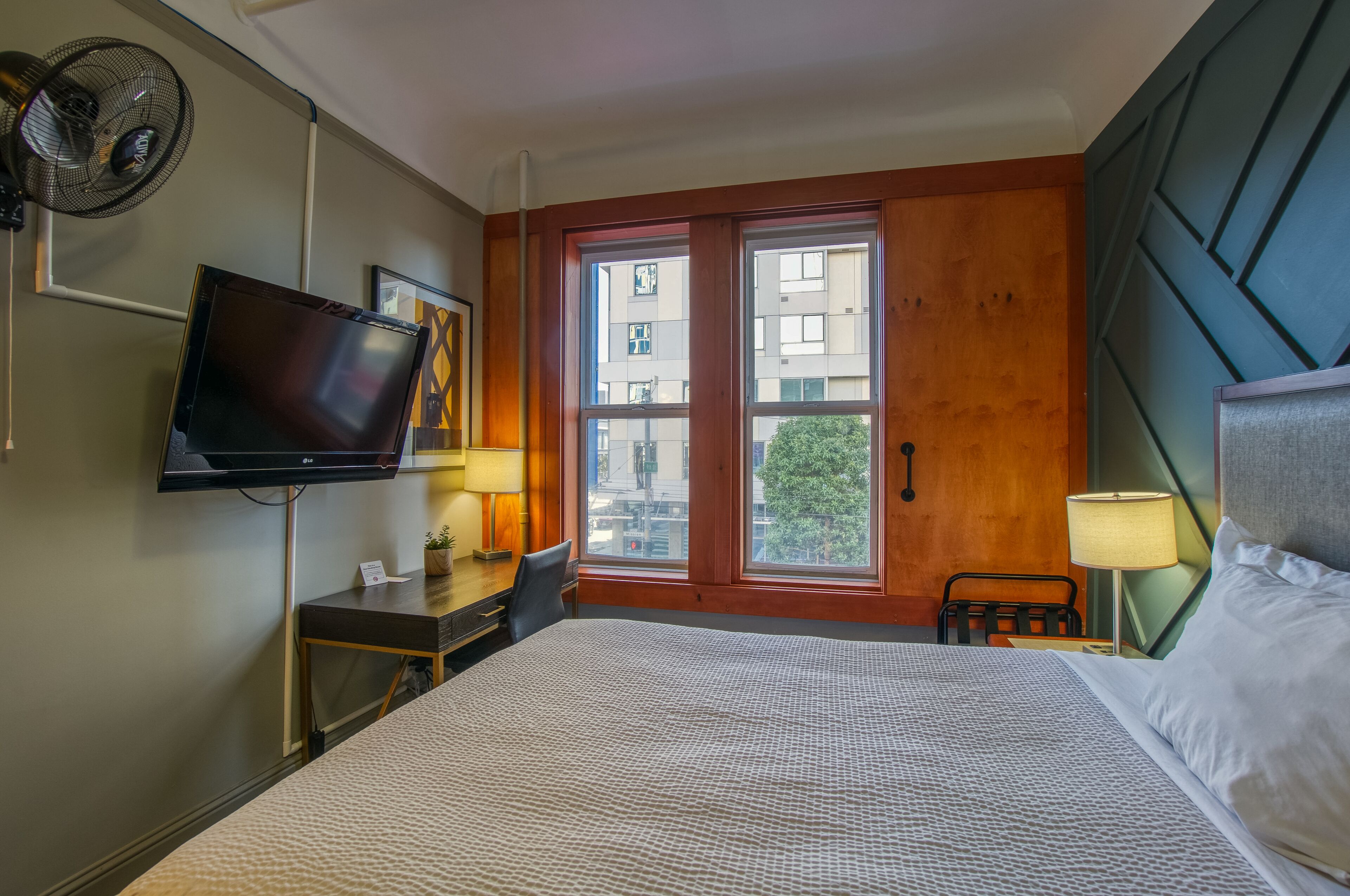 Habitación de hotel con una cama grande, un televisor montado en la pared, un escritorio con una silla y una ventana con vistas a edificios.