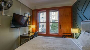 Habitación de hotel con una cama grande, un televisor montado en la pared, un escritorio con una silla y una ventana con vistas a edificios.