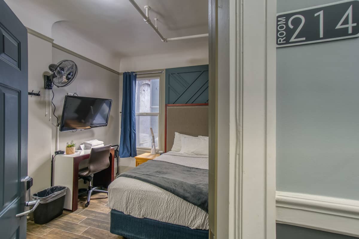 A hotel room with a bed, desk, chair, TV, and a trash bin.