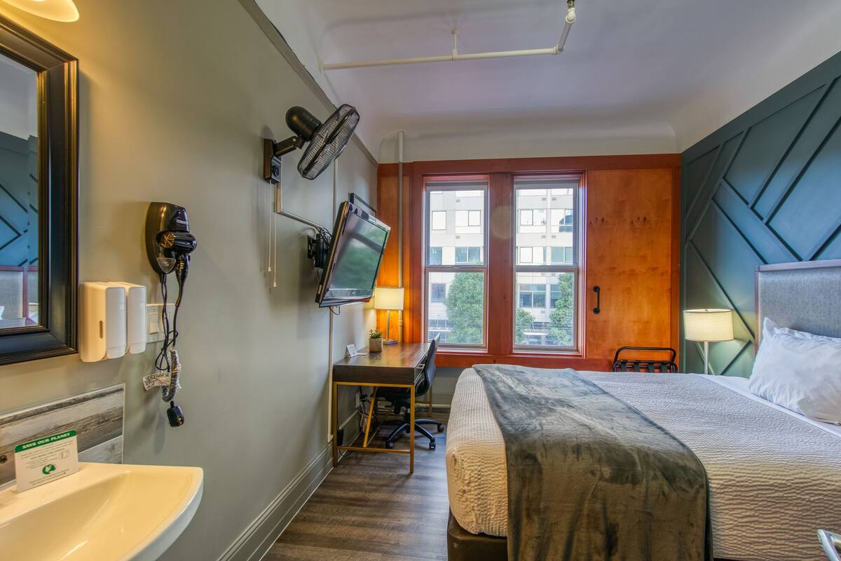 A hotel room with a bed, desk, chair, TV, and a window with a view of buildings.