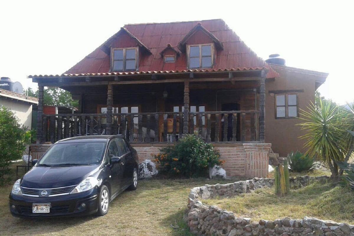 A two-story house with a red roof, a porch with wooden railings, and a stone wall in the front yard.