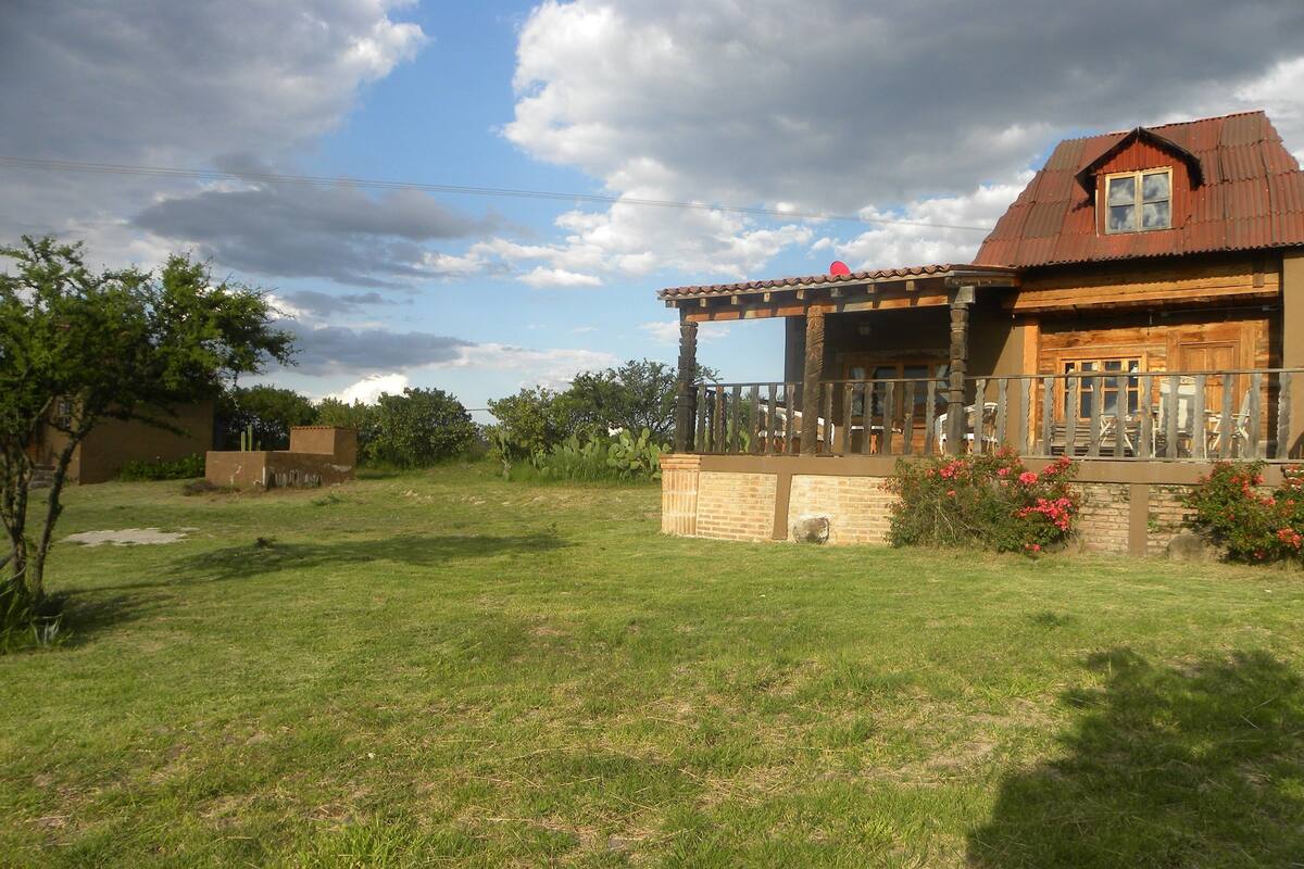 A rustic wooden house with a balcony, surrounded by a grassy yard and cacti.