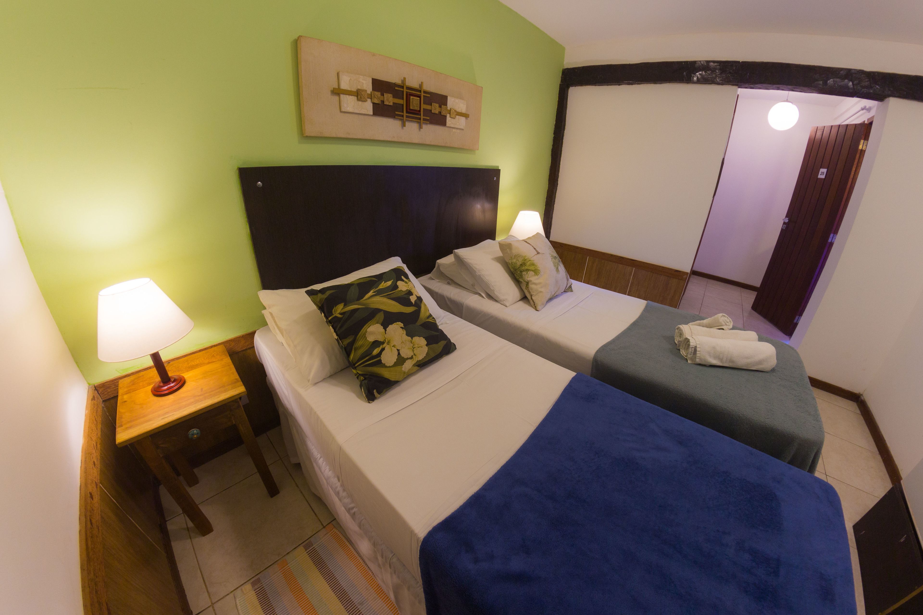 A hotel room with two beds, a nightstand with a lamp, a wall-mounted artwork, and a door leading to another room.