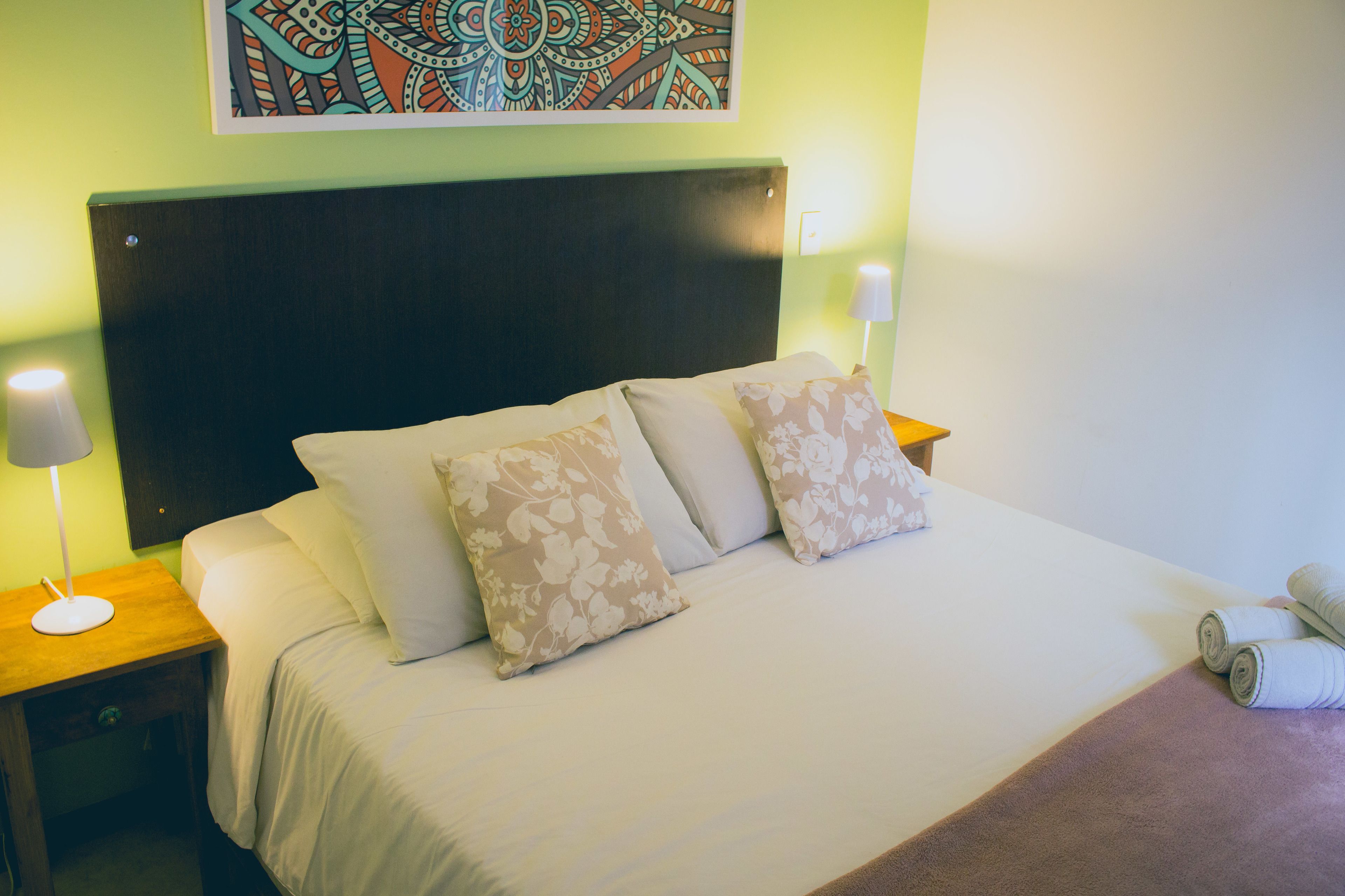 Cama bem arrumada com almofadas florais, mesa de cabeceira de madeira com abajur e uma obra de arte na parede acima da cama.