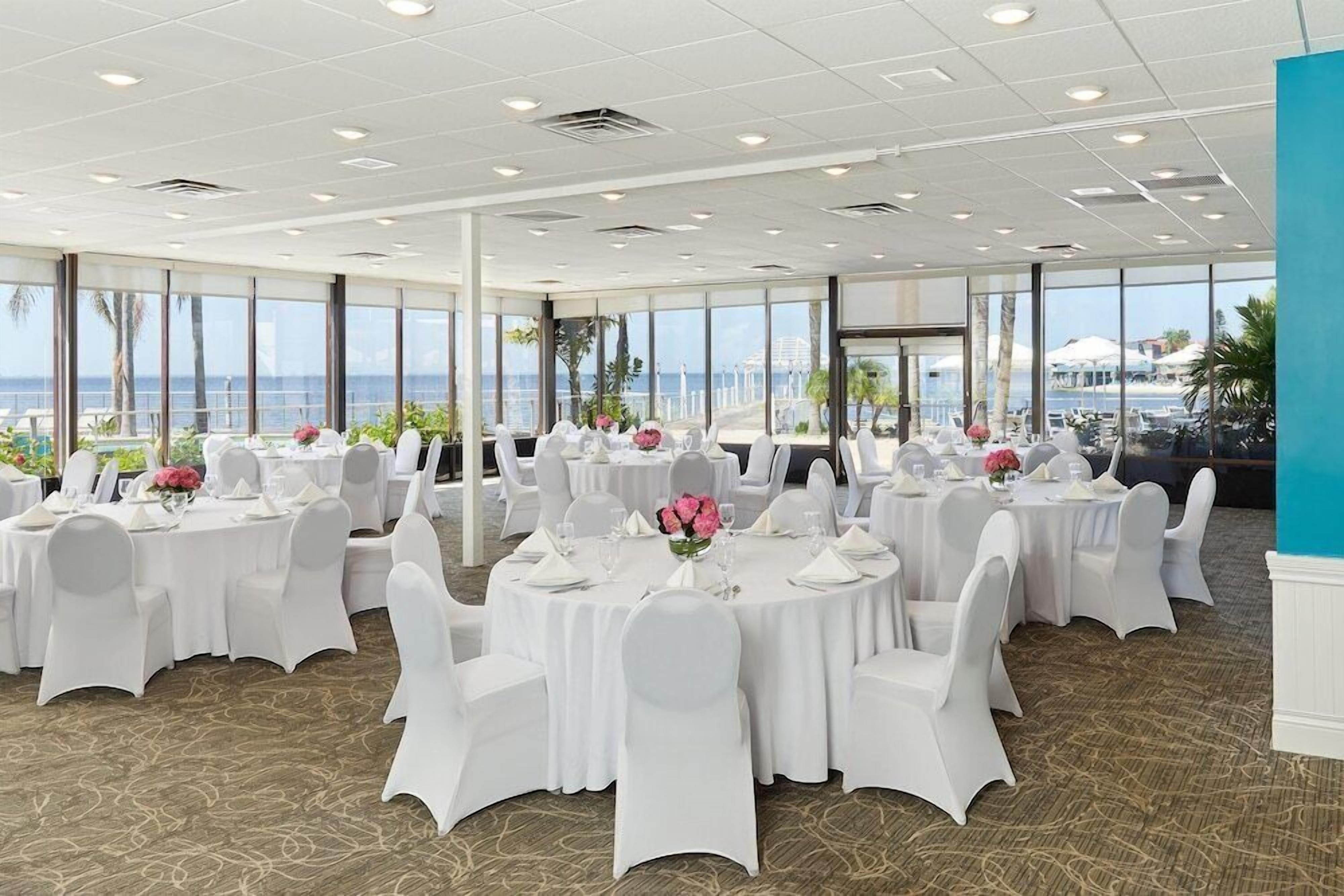 A banquet hall with round tables, white chairs, and large windows overlooking the sea.