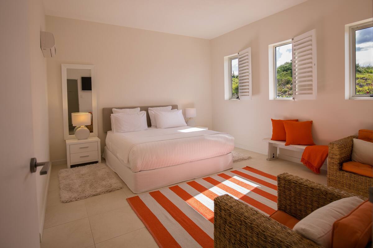 A bedroom with a bed, bedside tables, a mirror, a window with shutters, and a view of greenery outside.