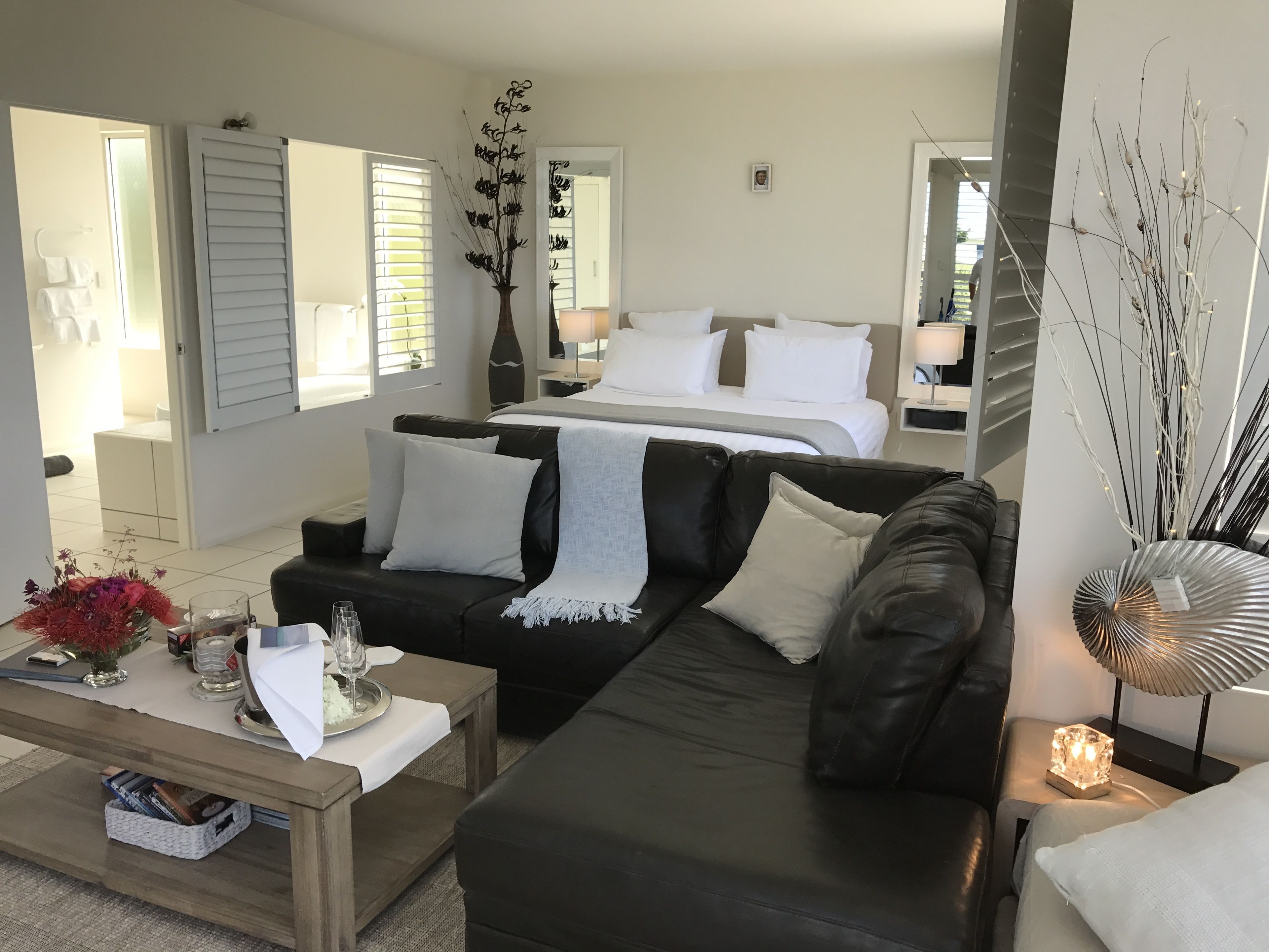 Una habitación de hotel moderna con un sofá de cuero negro, una mesa de centro de madera, una cama con ropa blanca y un espejo grande.