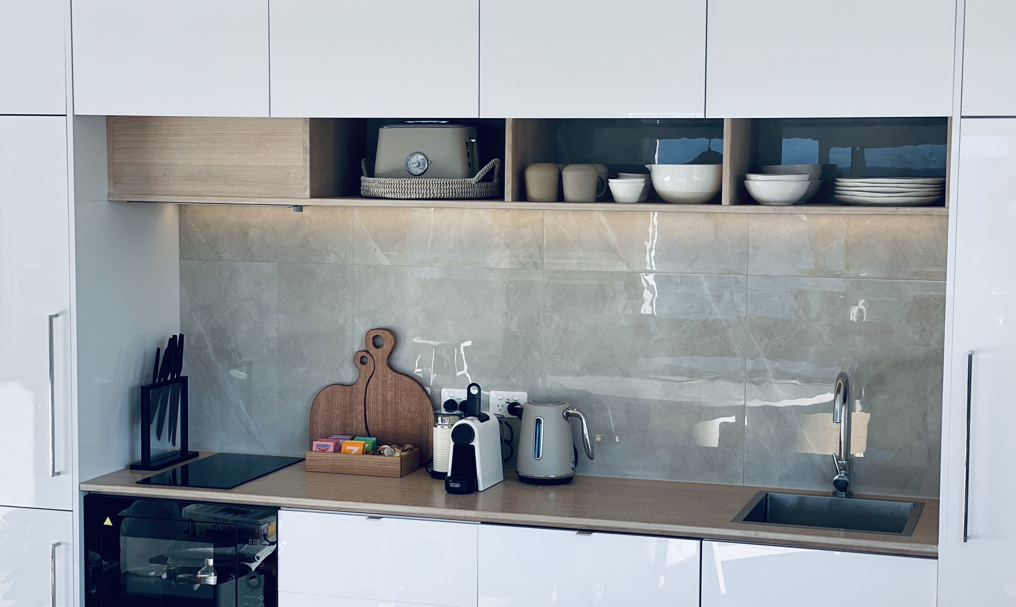 A modern kitchen with white cabinets, a wooden cutting board, a coffee maker, a kettle, and a sink.