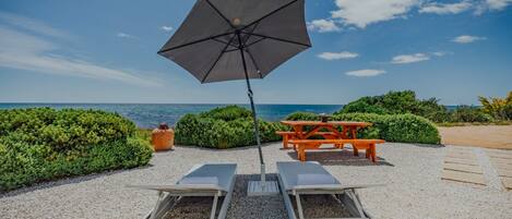 Eine Strandlounge mit großer Sonnenschirm, Liegestühlen, einem Picknicktisch und Blick auf das Meer.