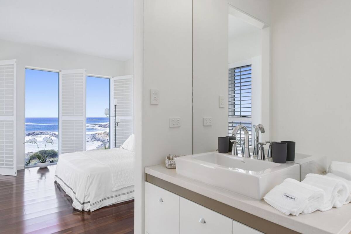 A modern hotel room with a large bed, a white bathroom with a sink and mirror, and a view of the ocean through open windows.
