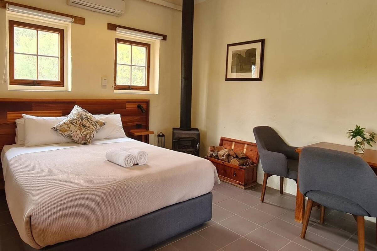 Une chambre à coucher avec un grand lit, deux fenêtres, une cheminée, un bureau avec deux chaises et un tableau encadré au mur.