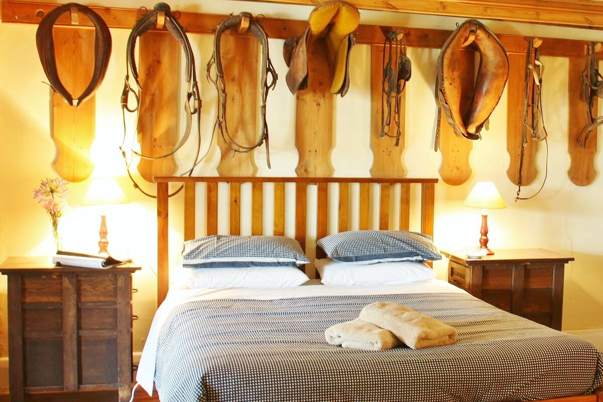 Un lit avec une couvre-lit à carreaux, deux oreillers et une serviette enroulée. On y trouve des têtes de lit en bois et un porte-selles fixé au mur.