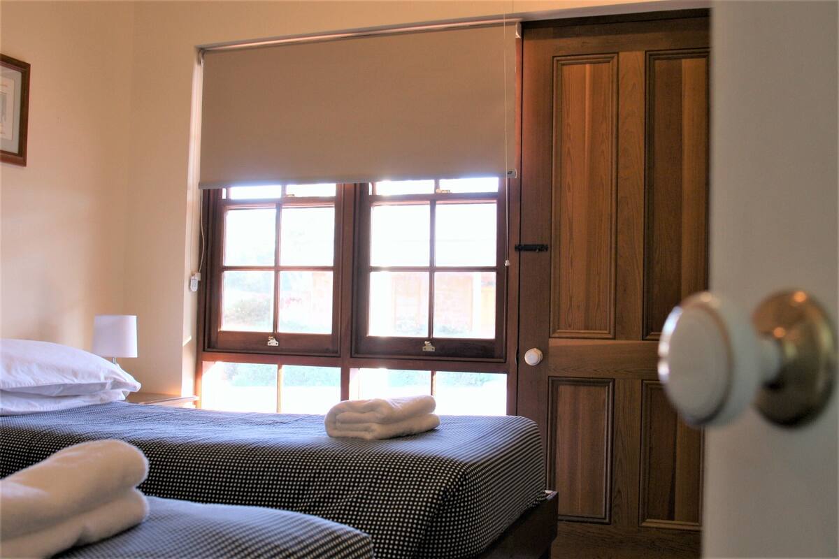 Une chambre d’hôtel avec deux lits, une fenêtre avec des stores, une porte en bois et une lampe de chevet.
