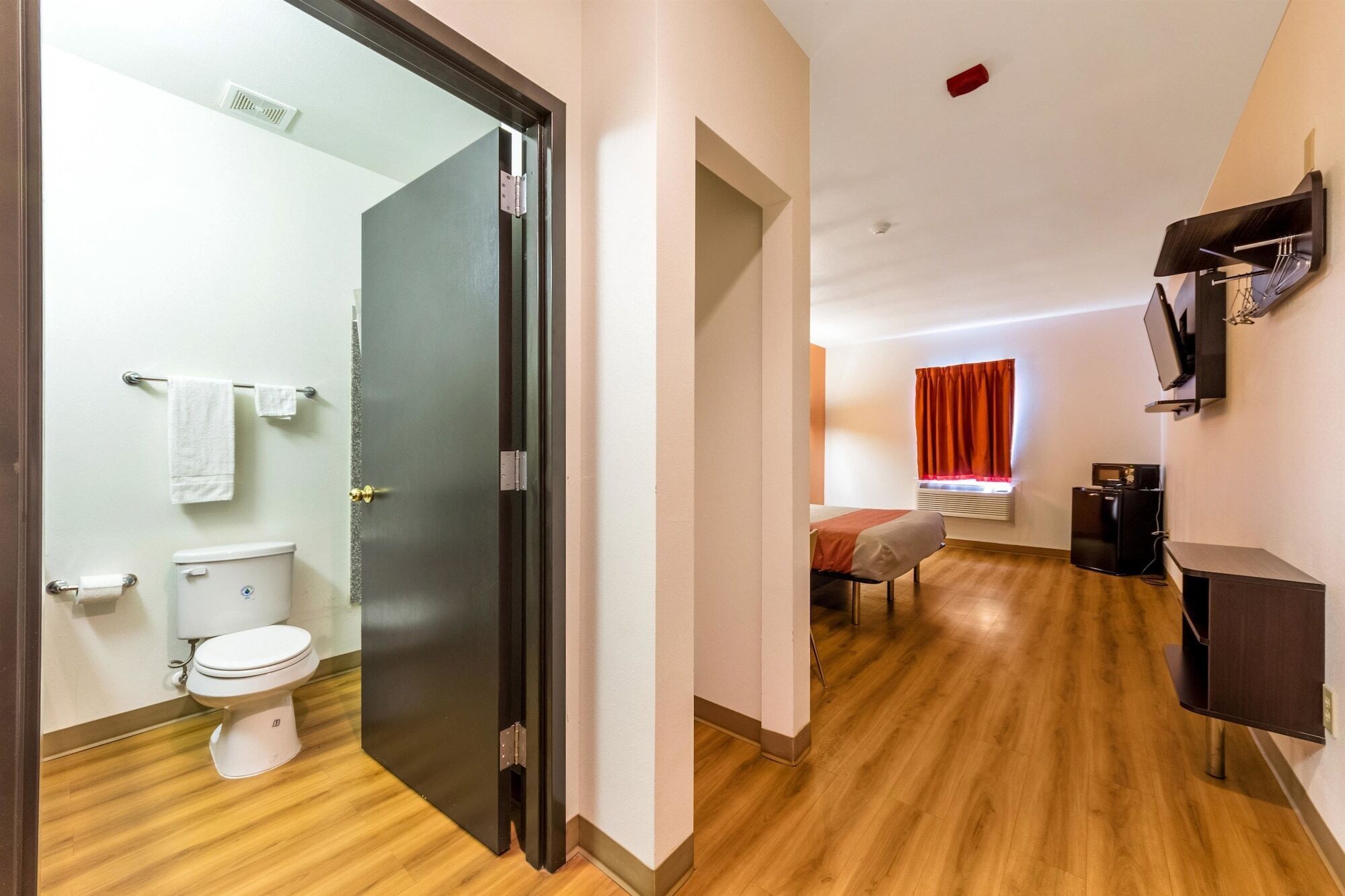 A modern hotel room with a wooden floor, a bathroom with a toilet and towel rack, a bed with a red curtain, a TV mounted on the wall, and a small kitchenette.