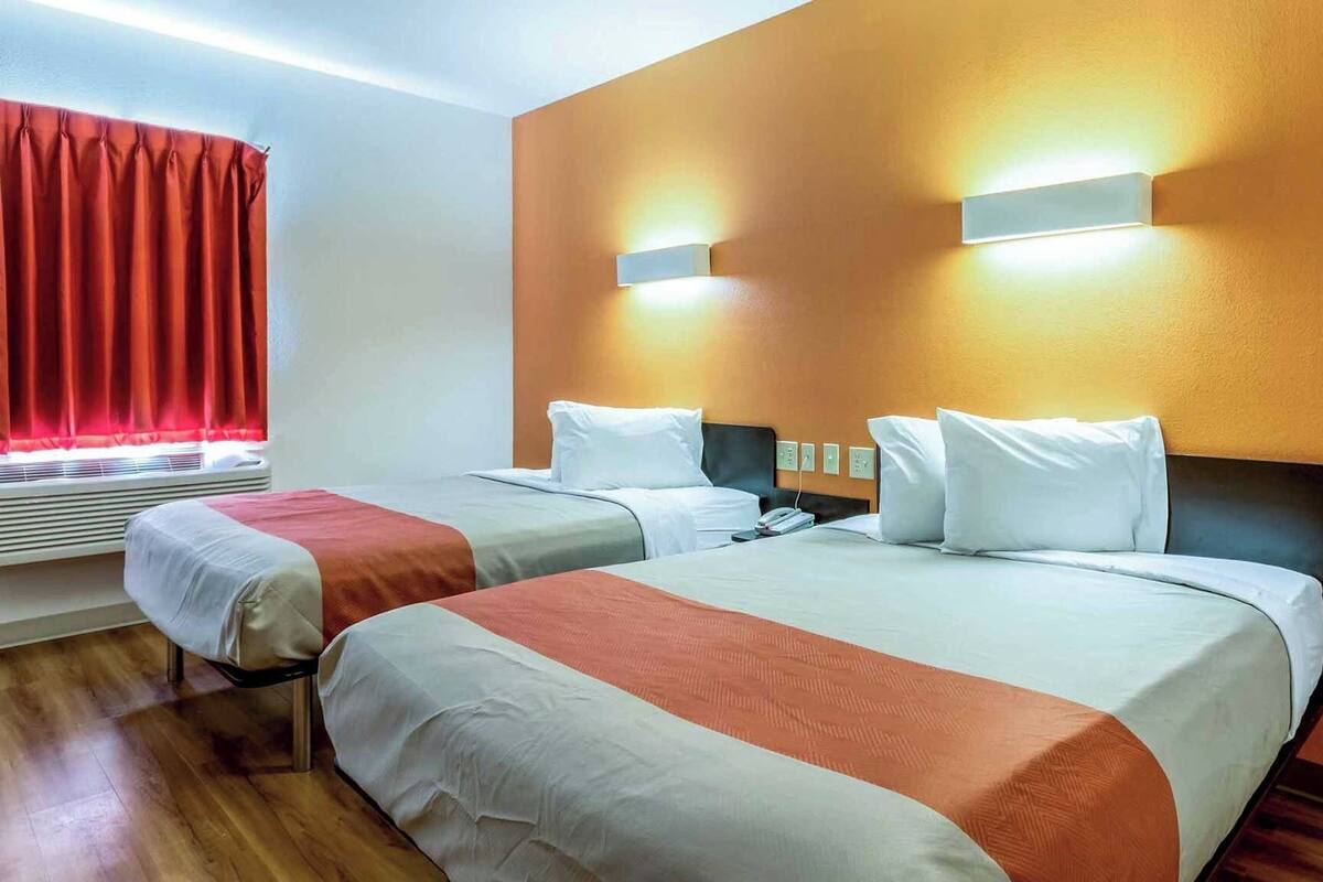 A hotel room with two beds, a window with red curtains, an air conditioning unit, and wall-mounted lights.