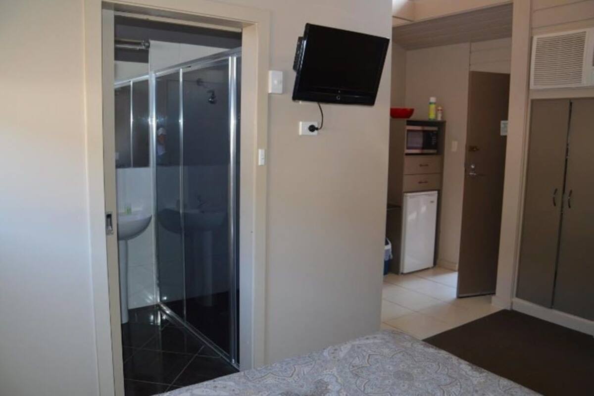A hotel room with a bed, a TV mounted on the wall, a shower, a sink, and a small kitchenette.