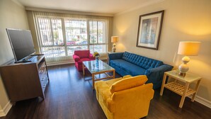 A living room with a blue sofa, a yellow armchair, a red chair, a glass coffee table, a TV on a wooden stand, and two lamps on side tables. - Cal Mar Hotel Suites (Santa Monica)