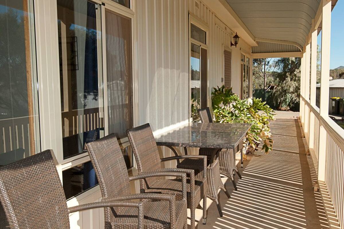 A covered outdoor patio with wicker chairs and a table, large windows, and a view of the surrounding landscape.