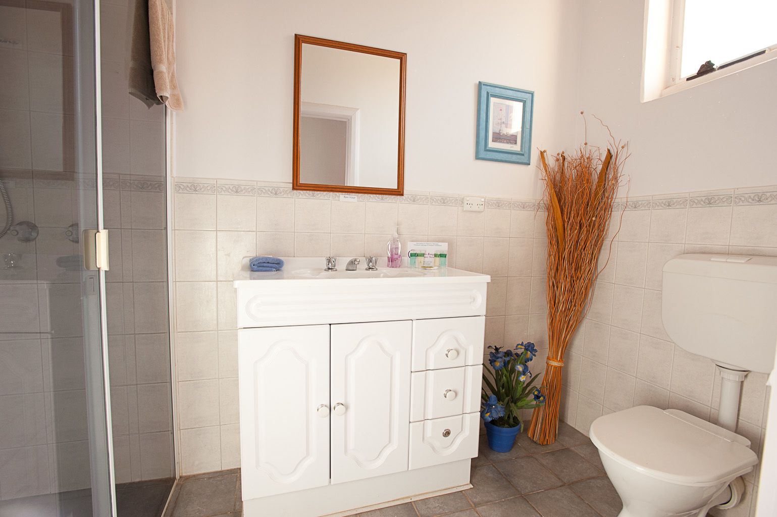 A bathroom with a white vanity, a mirror, a framed picture, a toilet, and a shower area.