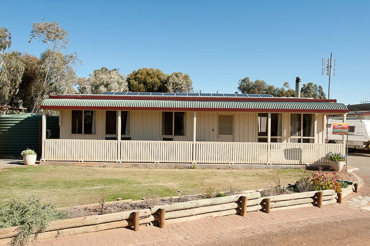 A small, single-story building with a green and white striped roof, a porch with white railings, and a sign indicating it is a 'Kangaroo Cottage'.