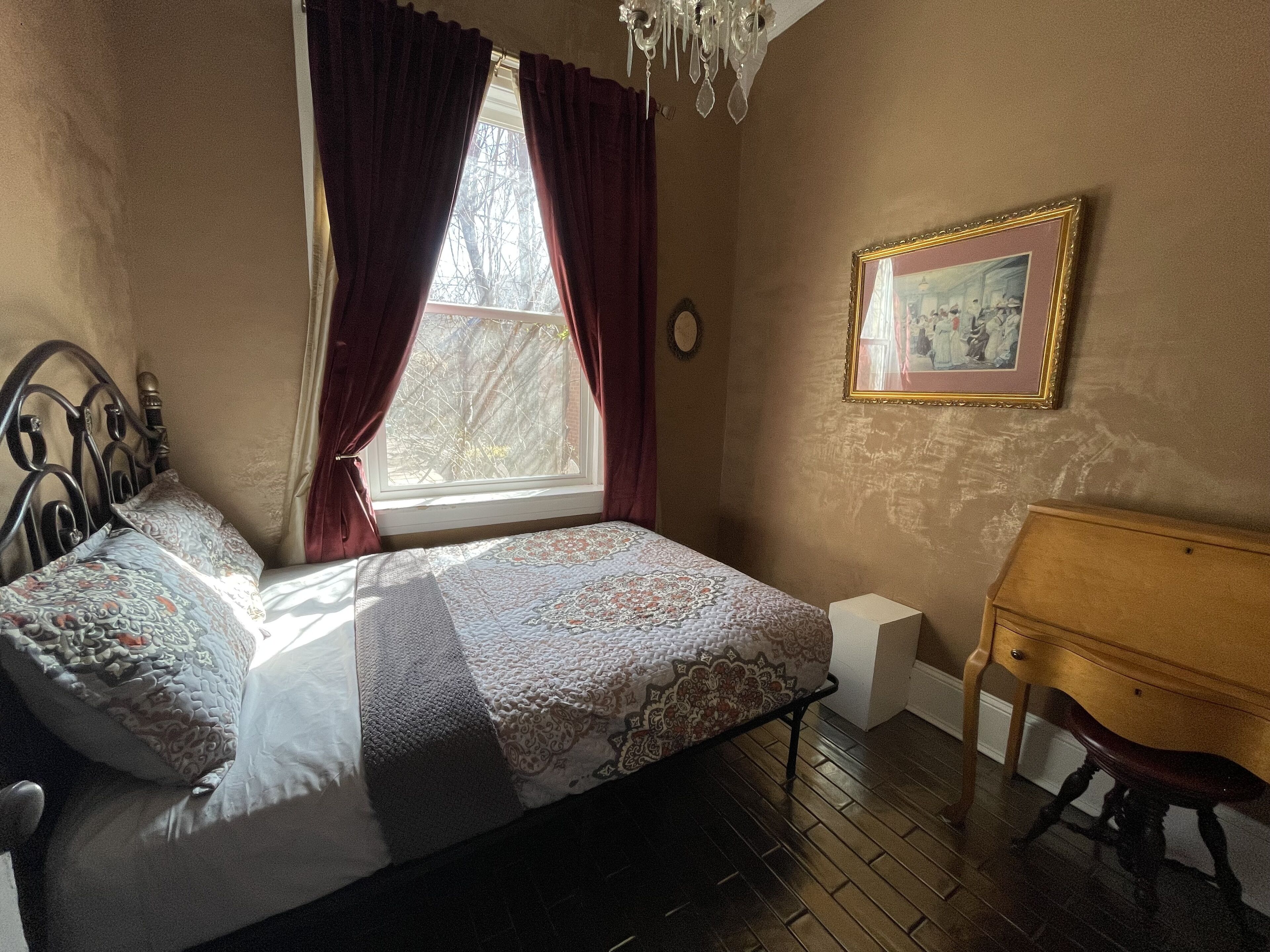A bedroom with a bed, a wooden bench, a window with curtains, and a framed picture on the wall.
