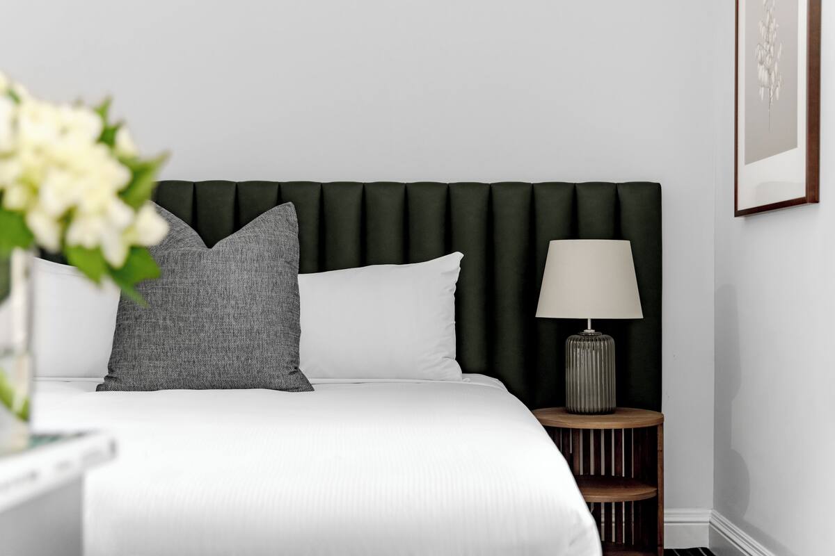 A neatly made bed with white and grey pillows, a dark green upholstered headboard, and a bedside table with a lamp.