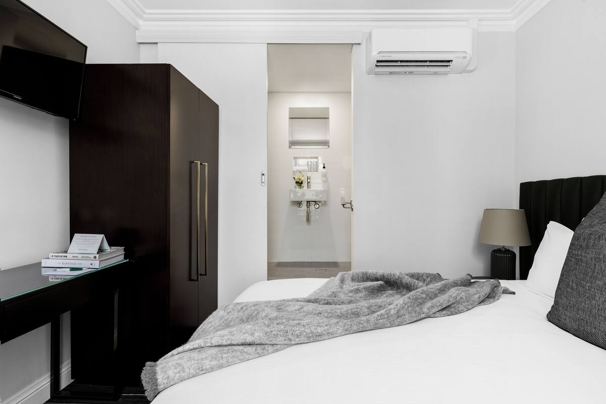 A hotel room with a bed, a desk with books, a TV, a wardrobe, a mirror, a bathroom with a sink and mirror, and an air conditioner.