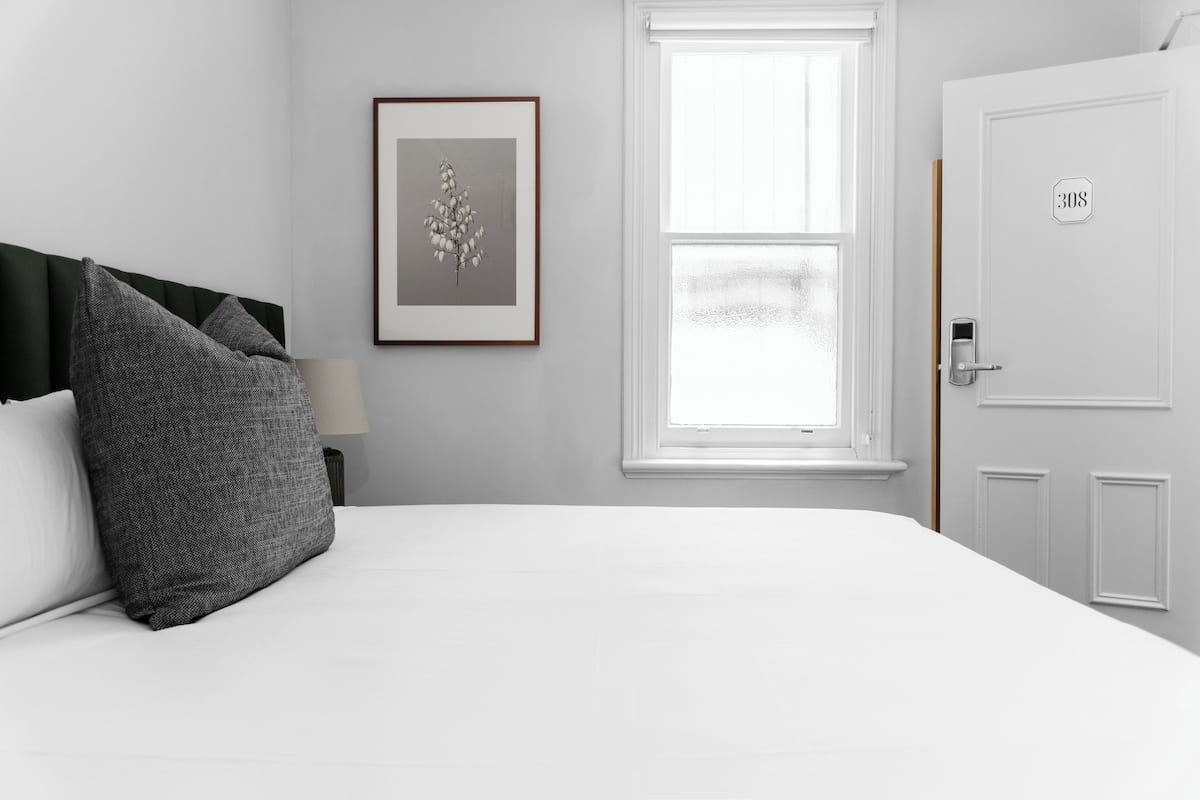 A neatly made bed with a white comforter and two pillows, a framed botanical print on the wall, a window with a view, and a door with a number plate.
