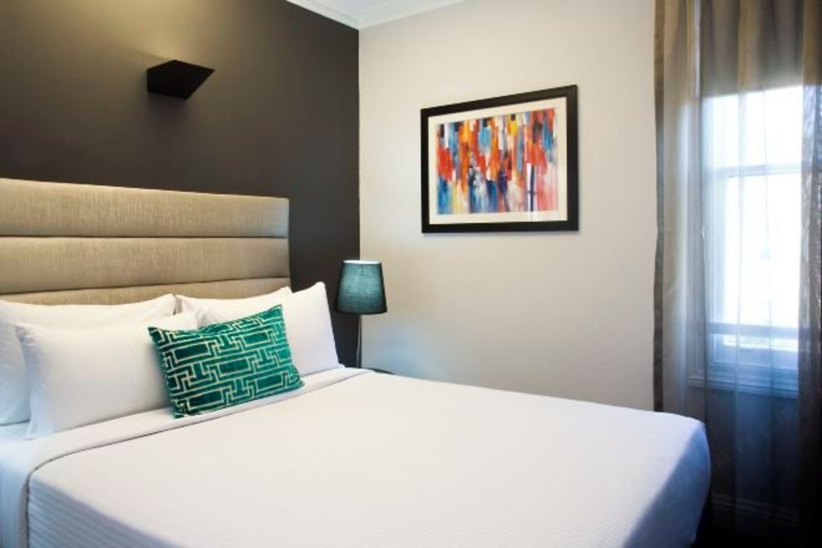 A hotel room with a large bed, a headboard, a wall-mounted lamp, and a framed abstract painting.