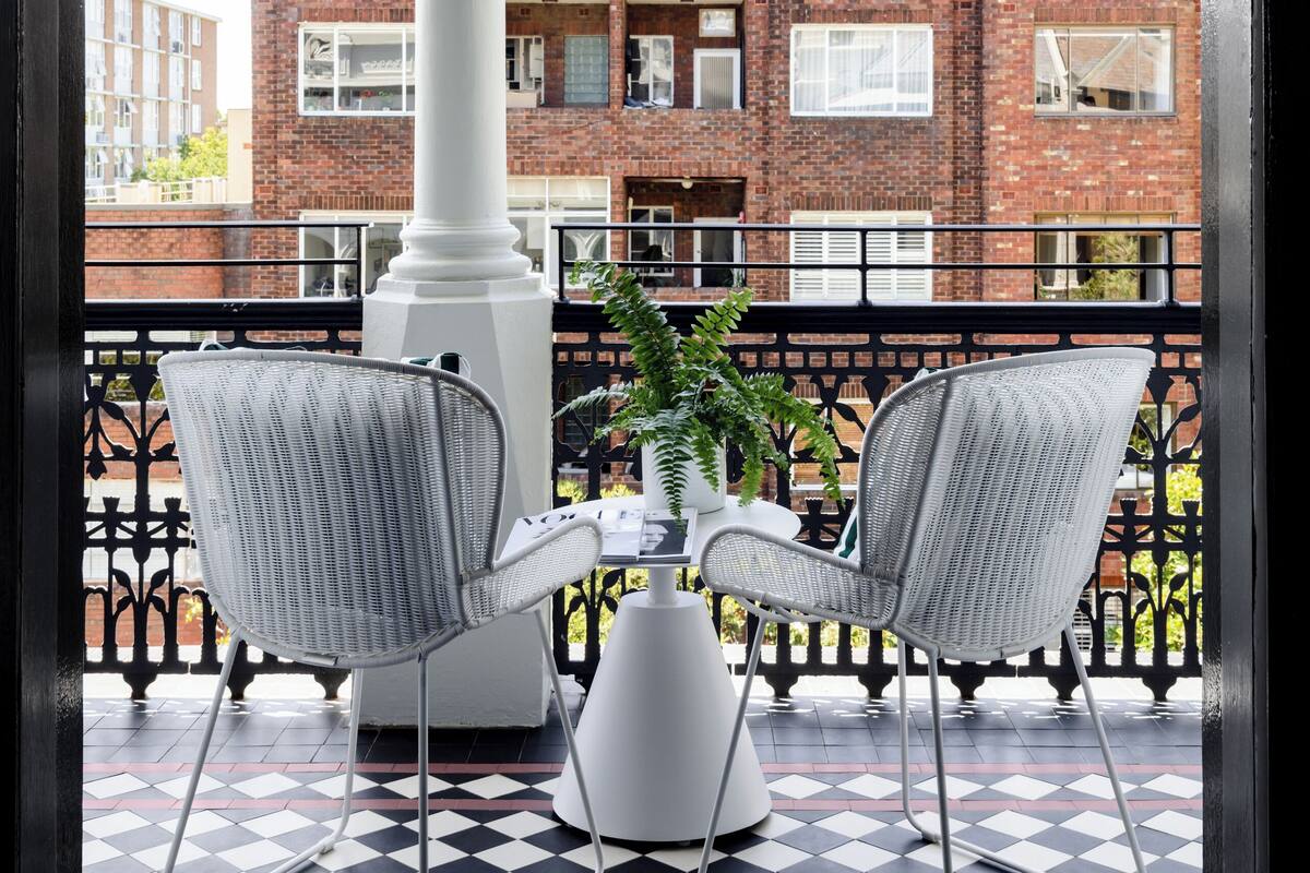 A balcony with two wicker chairs, a white table, and a potted plant. The balcony has a black and white checkered floor and a white railing. In the background, there is a brick building with windows and balconies.