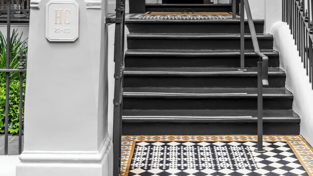 Una escalera conduce a la entrada de un edificio, con un felpudo de piso a cuadros blanco y negro, columnas blancas y un cartel que indica las habitaciones 21 al 23.