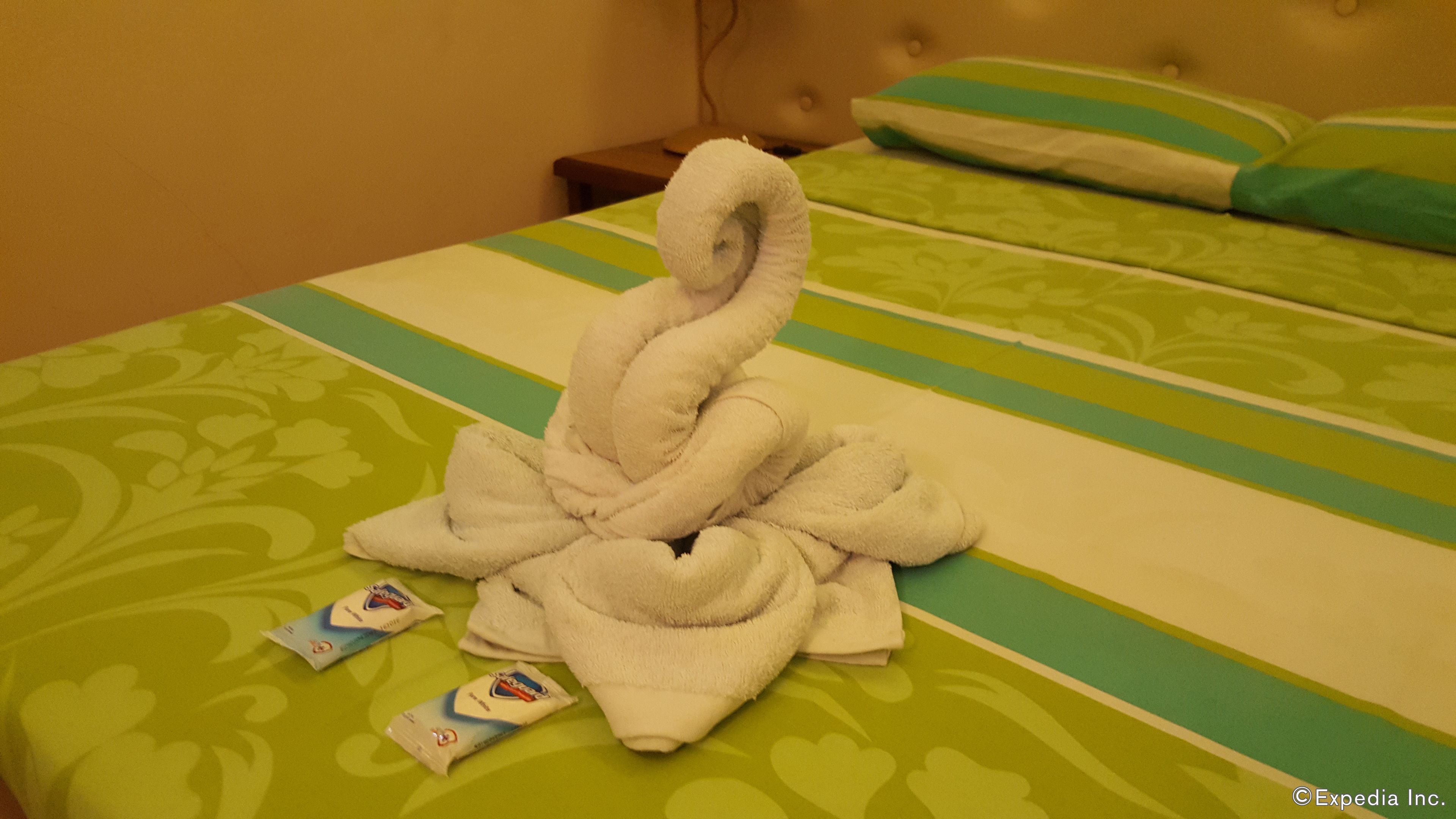 Habitación de hotel con cama, ropa de cama a rayas verdes y blancas, una toalla blanca con forma de cisne y dos pequeños sobres sobre la cama.