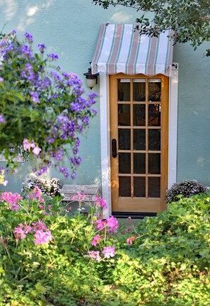 A door with a striped awning, surrounded by lush greenery and purple flowers. - Sandra D's Country Guest House (Fallbrook)