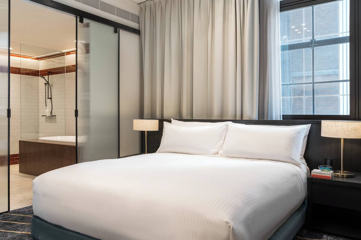 A hotel room with a large bed, a bedside table with a lamp, a window with curtains, and a view of the city.
