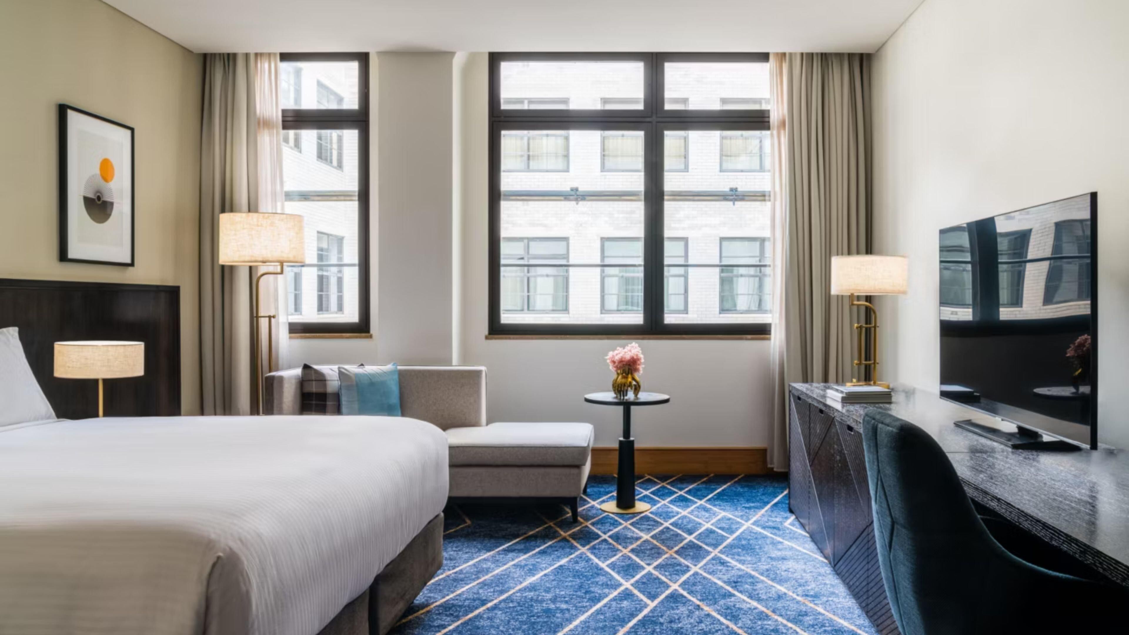 A hotel room with a bed, a desk, a chair, a small table with a vase, and a TV.